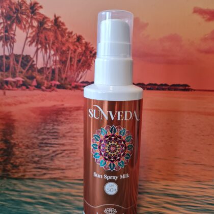 Sunveda Sun Spray Milk bottle held up in front of a tropical beach backdrop with palm trees and sunset. SPF 50+ label visible on the bottle.