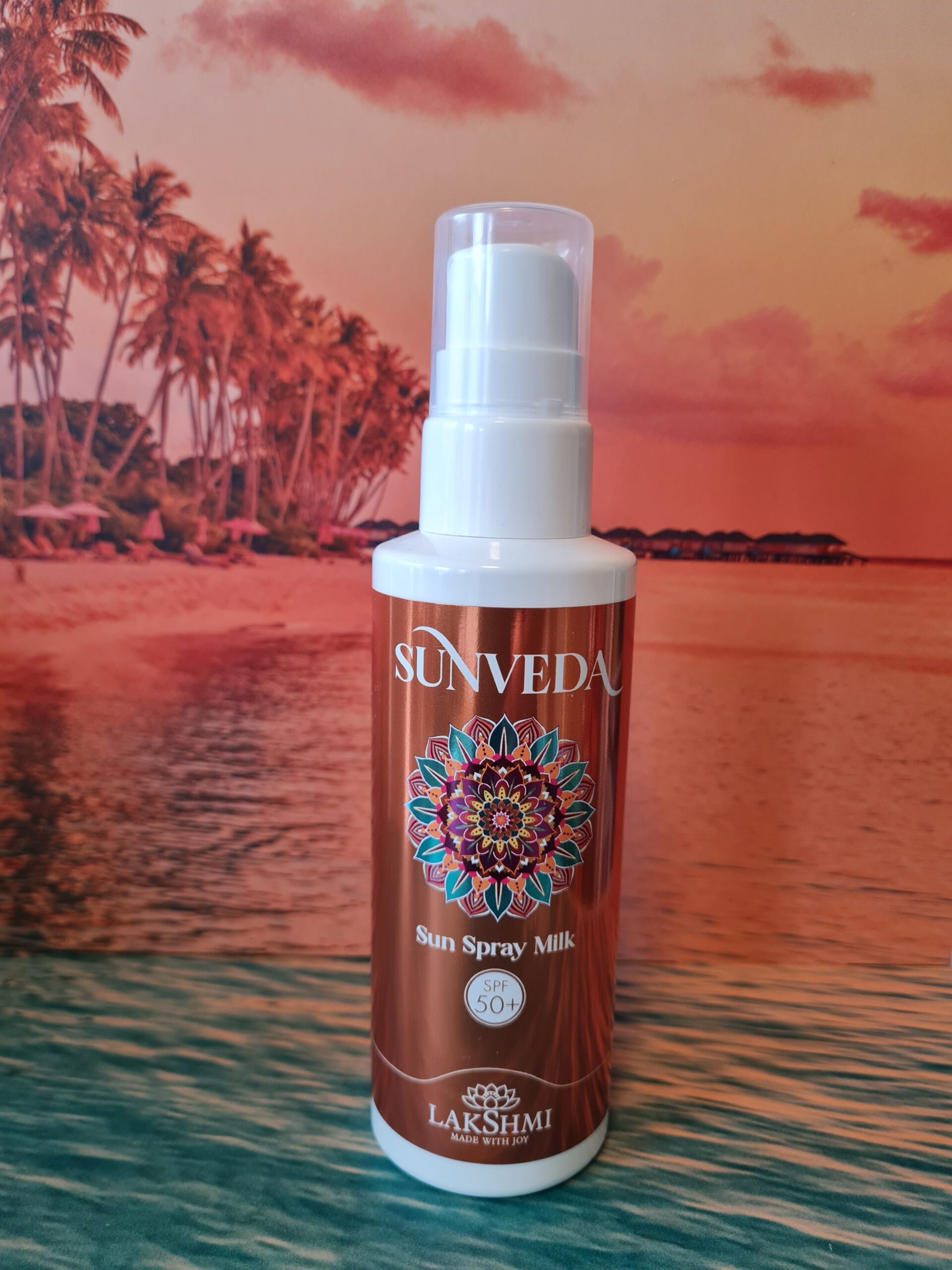 Sunveda Sun Spray Milk bottle held up in front of a tropical beach backdrop with palm trees and sunset. SPF 50+ label visible on the bottle.