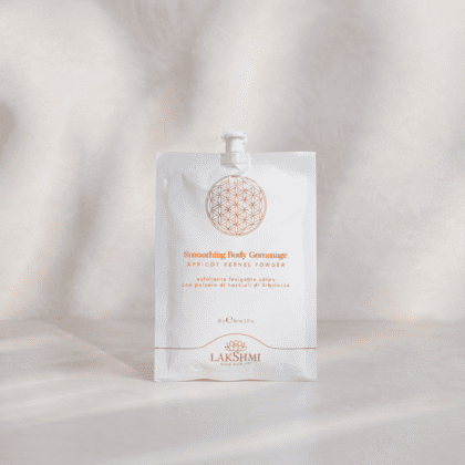 White cosmetic pouch with orange text and geometric logo, labeled 'Smoothing Body Gommage' with apricot kernel powder, Lakshmi brand.
